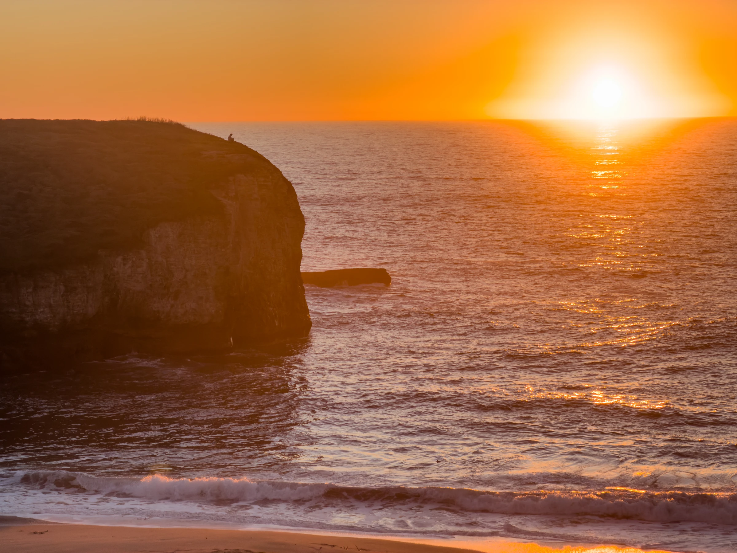 An orange sunset over the ocean, with a tiny figure on a sea cliff and waves rolling onto the beach.